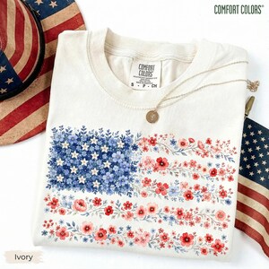 May include: An ivory t-shirt featuring a graphic of the American flag composed of blue, white, and red floral patterns. The shirt has a crew neckline and a Comfort Colors brand label at the inner collar. A gold-toned necklace rests on the fabric.