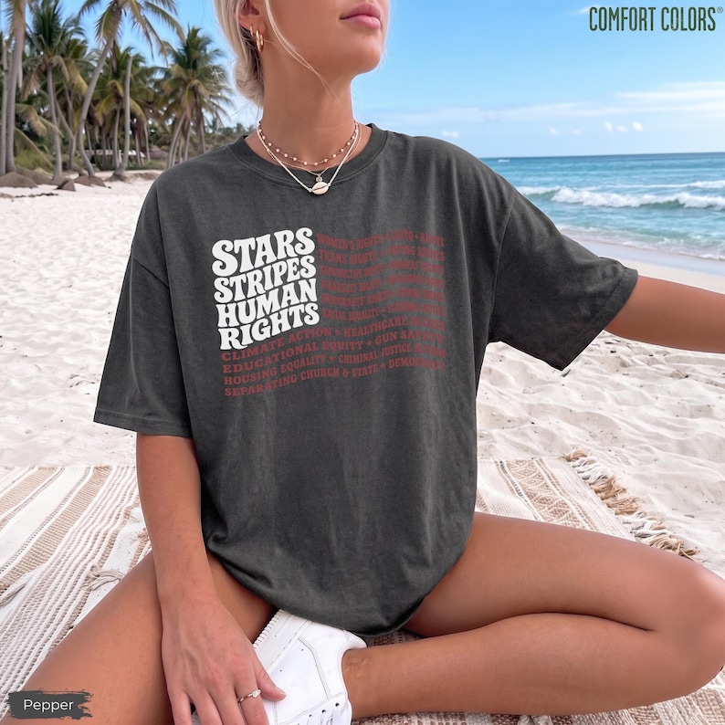May include: A charcoal gray short-sleeve t-shirt featuring white and red text that reads STARS STRIPES HUMAN RIGHTS, followed by a list of terms including CLIMATE ACTION, HEALTHCARE, EDUCATIONAL EQUITY, HOUSING EQUALITY, CRIMINAL JUSTICE, and DEMOCRACY.