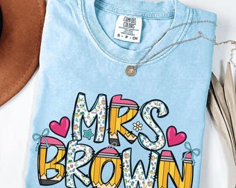 May include: A light blue short-sleeve t-shirt with a crew neckline and a Comfort Colors label. The graphic features the text "MRS BROWN" stylized with yellow pencil designs, colorful school-themed patterns, pink hearts, and small blue bows.
