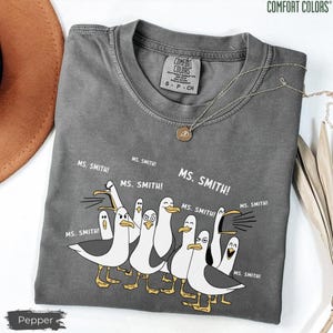 Puede incluir: Una camiseta gris Comfort Colors con un dise&ntilde;o de dibujos animados de gaviotas y el texto "MS. SMITH!". La camiseta tiene cuello redondo y mangas cortas. Un collar plateado con un colgante est&aacute; sobre la camiseta. La camiseta est&aacute; etiquetada "Pepper".