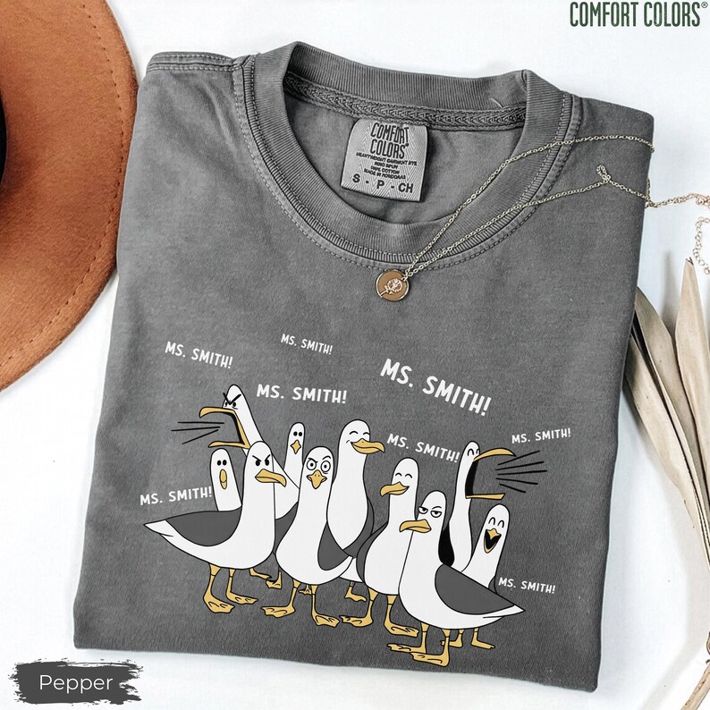 Puede incluir: Una camiseta gris Comfort Colors con un dise&ntilde;o de dibujos animados de gaviotas y el texto "MS. SMITH!". La camiseta tiene cuello redondo y mangas cortas. Un collar plateado con un colgante est&aacute; sobre la camiseta. La camiseta est&aacute; etiquetada "Pepper".