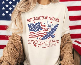 May include: Ivory-colored t-shirt with a patriotic design. The graphic includes the text "United States of America" above a waving American flag, the Statue of Liberty, and an eagle. The text "250th Anniversary 1776-2026" is below the flag.