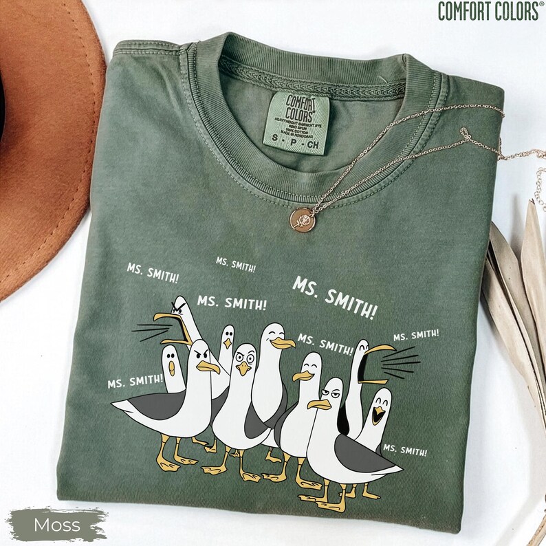 Puede incluir: Una camiseta verde musgo Comfort Colors con una caricatura de gaviotas y el texto "MS. SMITH!". La camiseta tiene cuello redondo y mangas cortas. Un collar dorado con un colgante est&aacute; drapeado sobre la camiseta.