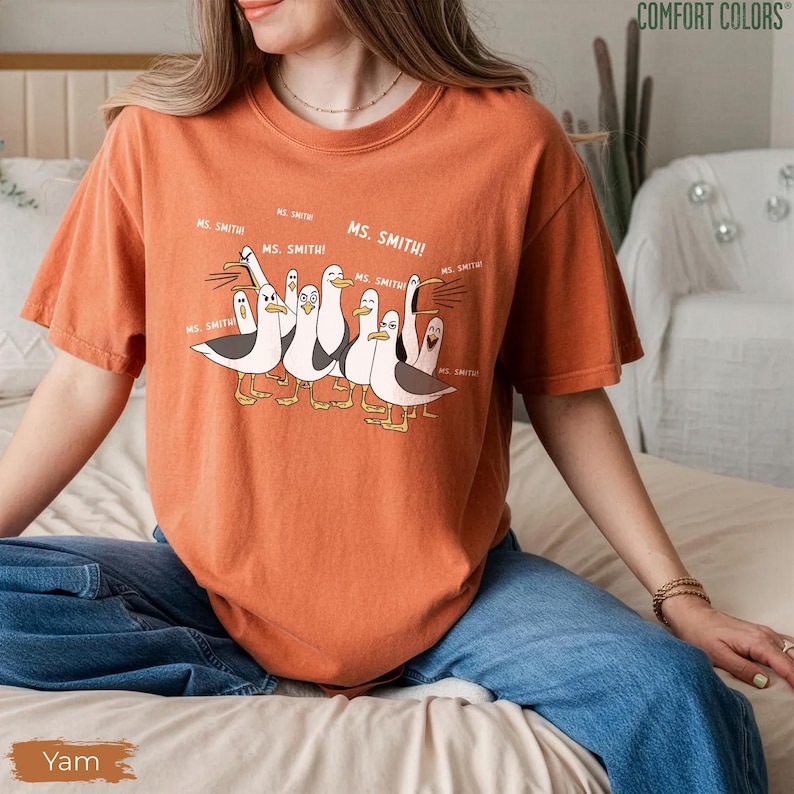 Puede incluir: Camiseta naranja Comfort Colors con una ilustraci&oacute;n de dibujos animados de gaviotas y el texto "MS. SMITH!" repetido. La camiseta tiene un corte holgado y mangas cortas. La palabra "Yam" est&aacute; impresa en la parte inferior izquierda.