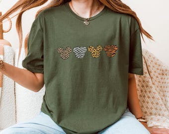 May include: Olive green t-shirt featuring four Mickey Mouse head shapes with animal print patterns: leopard, zebra, dalmatian, and tiger stripes. The shirt is a relaxed fit and has short sleeves. The text "Military Green" is visible in the bottom right corner.