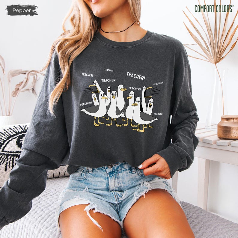 Puede incluir: Sudadera de manga larga gris oscuro con un gr&aacute;fico de gaviotas de dibujos animados, cada una con la palabra "TEACHER" encima. La sudadera es de la marca Comfort Colors. El color es "Pepper".