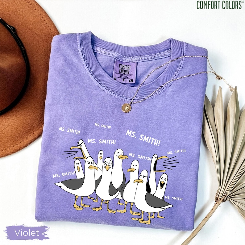 Puede incluir: Camiseta violeta Comfort Colors con una ilustraci&oacute;n de dibujos animados de gaviotas y el texto "MS. SMITH!". La camiseta tiene cuello redondo y mangas cortas. Un collar dorado con un colgante est&aacute; sobre la camiseta.