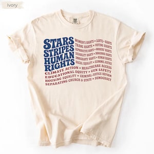 May include: An ivory short-sleeve t-shirt featuring a graphic design in blue and red text. The text reads Stars, Stripes, Human Rights, followed by a list of social causes including Women's Rights, LGBTQ+ Rights, and others in a wavy flag pattern.