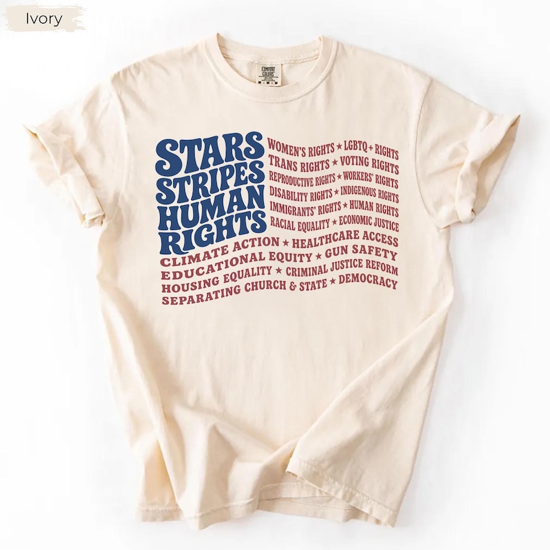 May include: An ivory short-sleeve t-shirt featuring a graphic design in blue and red text. The text reads Stars, Stripes, Human Rights, followed by a list of social causes including Women's Rights, LGBTQ+ Rights, and others in a wavy flag pattern.