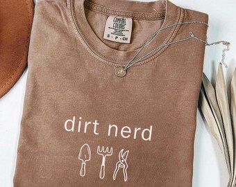 May include: Espresso-colored Comfort Colors t-shirt with the words "dirt nerd" and gardening tool graphics. A silver necklace with a pendant rests on the shirt. The shirt is size small.