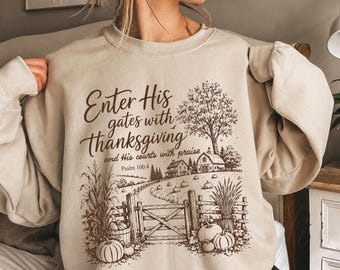 May include: A sand-colored crewneck sweatshirt featuring a brown graphic print of a farm scene with a wooden gate, pumpkins, corn stalks, and a tree. The text reads: Enter His gates with thanksgiving and His courts with praise. Psalm 100:4.