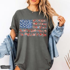 May include: A charcoal gray short-sleeve t-shirt featuring a graphic print of the American flag composed entirely of small blue, white, and red flowers. The shirt has a crew neckline and a loose fit, with the word Pepper printed in the corner.