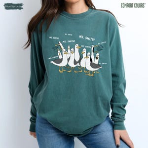 Puede incluir: Una sudadera Comfort Colors de color verde azulado con una ilustraci&oacute;n de dibujos animados de gaviotas. Las gaviotas est&aacute;n en grupo, con el texto "MS. SMITH!" encima. La sudadera es de color liso con cuello redondo.
