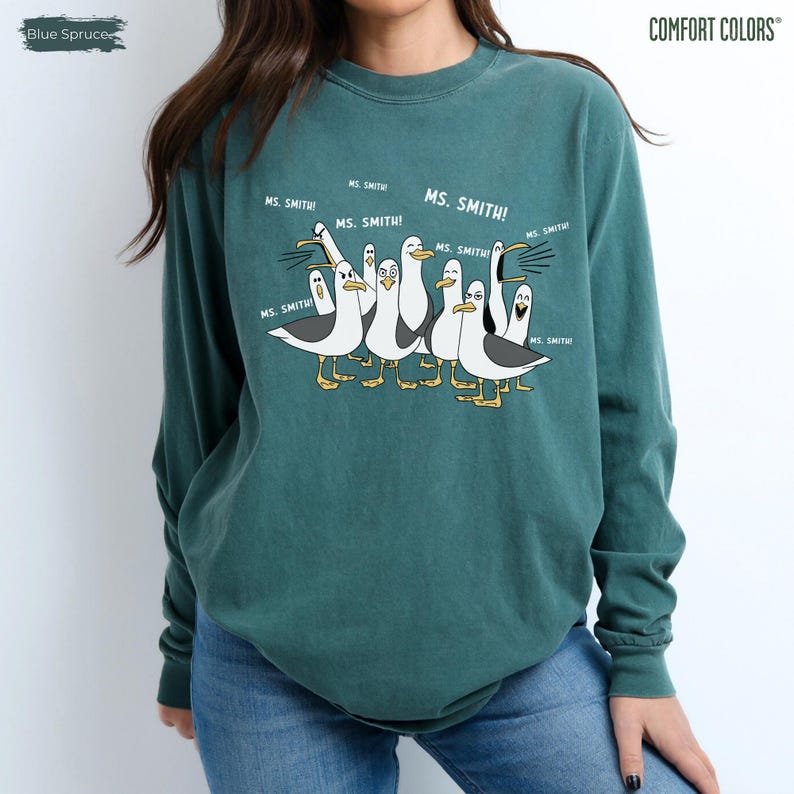 Puede incluir: Una sudadera Comfort Colors de color verde azulado con una ilustraci&oacute;n de dibujos animados de gaviotas. Las gaviotas est&aacute;n en grupo, con el texto "MS. SMITH!" encima. La sudadera es de color liso con cuello redondo.