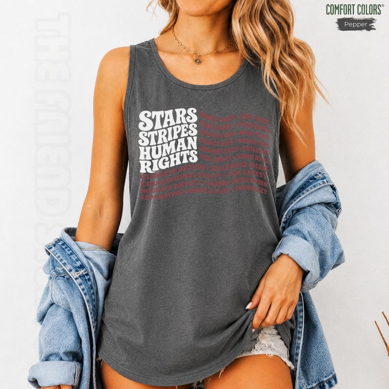 May include: A charcoal gray sleeveless tank top featuring the white text STARS STRIPES HUMAN RIGHTS arranged in a wavy flag pattern. The garment is worn with a light blue denim jacket draped over the shoulders. The brand label reads COMFORT COLORS Pepper.
