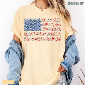 May include: A pale yellow short-sleeve t-shirt featuring a graphic of the American flag composed entirely of small blue, red, and white flowers. The shirt has a crew neckline and is layered under a light blue denim jacket. The text COMFORT COLORS is visible.