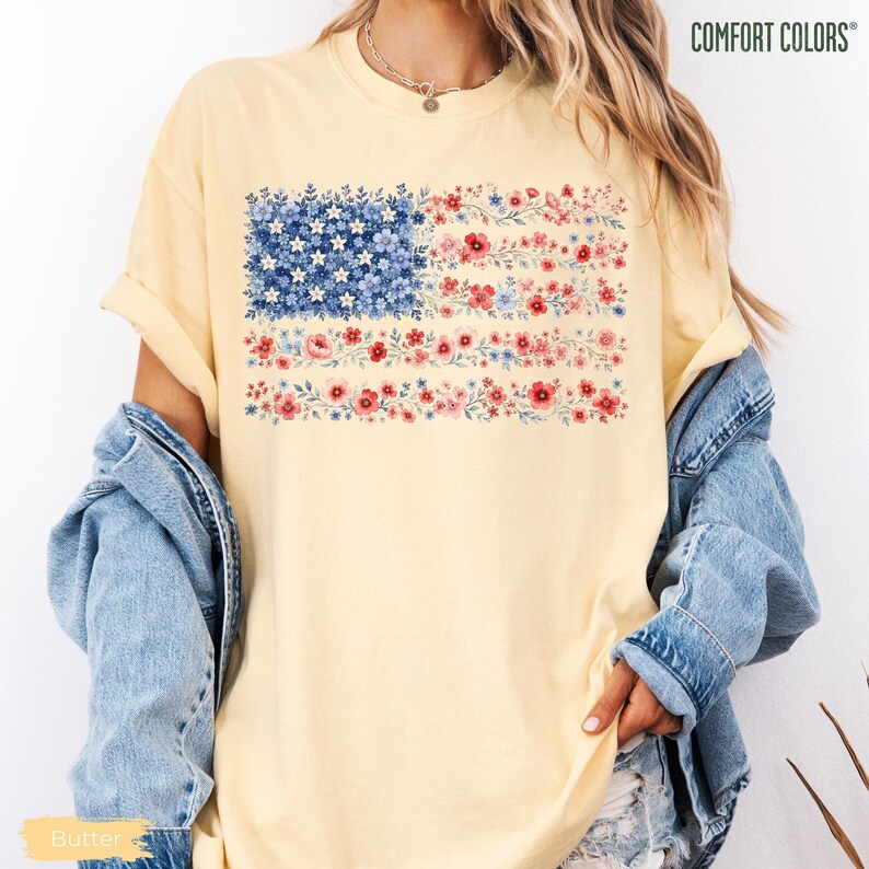 May include: A pale yellow short-sleeve t-shirt featuring a graphic of the American flag composed entirely of small blue, red, and white flowers. The shirt has a crew neckline and is layered under a light blue denim jacket. The text COMFORT COLORS is visible.