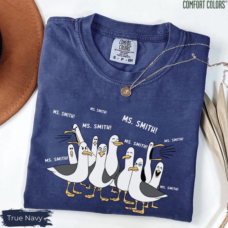 Puede incluir: Una camiseta azul marino con un gr&aacute;fico de gaviotas de dibujos animados y el texto "MS. SMITH!". La camiseta de la marca Comfort Colors es de talla S-P-CH. Un collar dorado con un colgante est&aacute; sobre la camiseta.