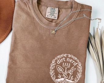 May include: Espresso-colored t-shirt with the words "dirt nerd" in a white circular design. The shirt has a crew neck and short sleeves. A silver necklace with a small pendant rests on the shirt.