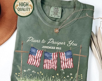 May include: A moss green Comfort Colors t-shirt with the text "Plans to Prosper You" and "Jeremiah 29:11". The shirt features three American flags hanging on a clothesline with a floral design at the bottom. A gold necklace is draped on the shirt.