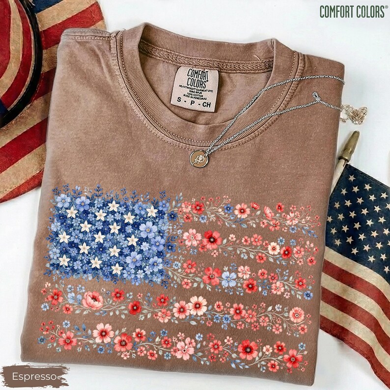 May include: A brown short-sleeve t-shirt featuring a graphic of the American flag composed entirely of small blue, white, and red flowers. The shirt has a crew neckline with a Comfort Colors brand label and a silver chain necklace resting on it.