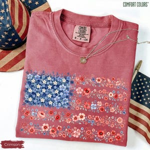 May include: A crimson t-shirt featuring a floral graphic arranged in the pattern of the American flag, with blue flowers forming the canton and red and white flowers forming the stripes. A silver necklace rests on the neckline of the shirt.