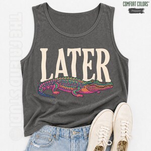 May include: A charcoal gray tank top featuring the word LATER in large cream-colored letters above a colorful graphic of an alligator. The alligator is illustrated with vibrant shades of green, pink, and purple. The garment has a crew neckline.
