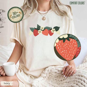 May include: Ivory-colored t-shirt featuring a strawberry design with red strawberries, green leaves, and white flowers. The shirt has the text "30+ COLORS" in a circular graphic and "COMFORT COLORS" in the upper right corner.