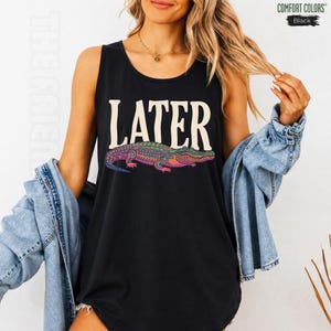 May include: A black sleeveless tank top featuring the word LATER in large cream-colored letters above a colorful, patterned alligator graphic. The design includes a Comfort Colors brand label in the top right corner and a light blue denim jacket.