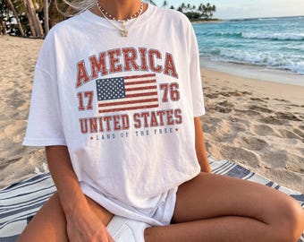 May include: White t-shirt with a distressed graphic featuring the word "AMERICA", the American flag, and the text "UNITED STATES LAND OF THE FREE". The shirt is a relaxed fit.