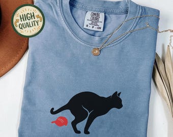 May include: Blue jean colored t-shirt with a black cat graphic and a red hat. The shirt has the words "Comfort Colors" printed on the top. A gold necklace is draped across the shirt.