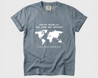 May include: A dusty blue t-shirt with rolled-up sleeves. The shirt features a white world map graphic and the text "DEAR WORLD, WE ARE SO SORRY. LOVE, SANE AMERICANS."