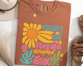 Op de afbeelding: Een yam-kleurig Comfort Colors t-shirt met de tekst "SCHOOL'S OUT FOREVER RETIREMENT" in een kleurrijk bloemenontwerp. Het shirt is gestyled met een gouden ketting, een hoed en sandalen.
