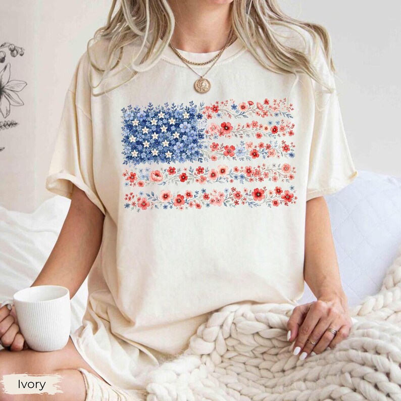 May include: An ivory short-sleeve t-shirt featuring a graphic print of the American flag composed entirely of small blue, white, and red flowers. The flag design is rectangular, with a blue field of flowers on the left and horizontal stripes of red and white floral patterns extending to the right.