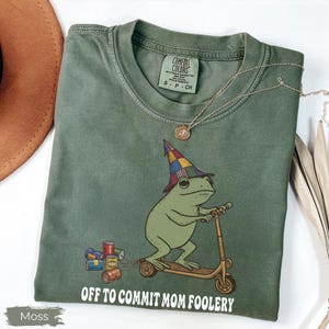 May include: Moss green t-shirt with a cartoon frog on a scooter, wearing a colourful wizard hat. The text "OFF TO COMMIT MOM FOOLERY" is printed below the frog. A gold necklace is draped on the shirt.