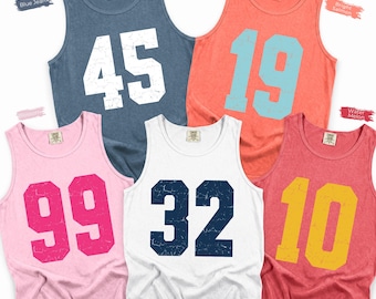 May include: Five sleeveless tank tops in various colors: blue jean with white 45, bright salmon with light blue 19, blossom with hot pink 99, white with navy blue 32, and watermelon with yellow 10. Each shirt features a distressed varsity number.