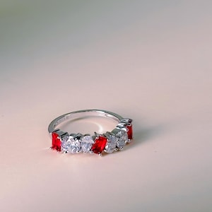 Könnte beinhalten: Ein silberner Ring mit abwechselnd roten und klaren Edelsteinen. Der Ring ist vor einem weichen, neutralen Hintergrund mit einem Zweig weißer Blumen in der oberen linken Ecke platziert. Die Edelsteine sind rechteckig.