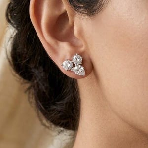 Può includere: Un orecchino a lobo color argento con un gruppo di tre pietre trasparenti sfaccettate. Il design include due pietre a taglio rotondo posizionate sopra una singola pietra a forma di pera, incastonate in una montatura metallica.