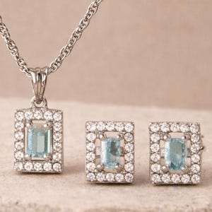 Könnte beinhalten: Eine silberne Halskette mit rechteckigem Anhänger und passenden Ohrringen. Jedes Schmuckstück hat einen hellblauen, rechteckigen Stein, der von kleinen, klaren Steinen umgeben ist. Das Schmuckset wird auf einer hellen Oberfläche präsentiert.