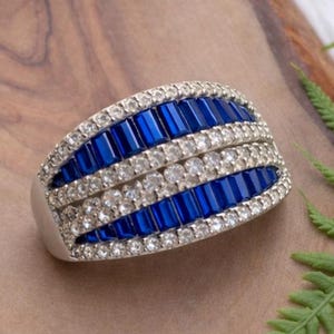 Könnte beinhalten: Ein silberner Ring mit einem rechteckigen Design mit abwechselnden Reihen aus blauen und klaren Edelsteinen. Der Ring liegt auf einer Holzoberfläche mit grünen Blättern.