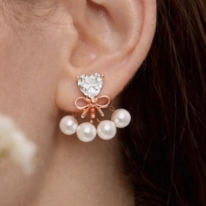 Könnte beinhalten: Ohrringe aus Roségold mit einem herzförmigen, klaren Edelstein und einer Schleife. Die Ohrringe sind mit drei weißen Perlen verziert. Ein modisches Accessoire.