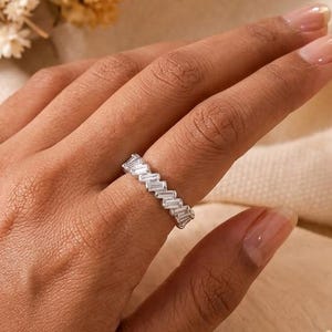 Könnte beinhalten: Ein silberner Ring mit Zickzack-Muster, besetzt mit rechteckigen, klaren Edelsteinen. Der Ring befindet sich an einer Hand mit gepflegten Nägeln, umgeben von Trockenblumen.