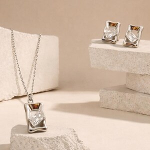 Könnte beinhalten: Eine silberfarbene Halskette mit Anhänger und passenden Ohrringen. Der Anhänger und die Ohrringe haben ein rechteckiges Design mit einem klaren, facettierten Stein und einem goldfarbenen Akzent. Der Schmuck wird vor neutralem Hintergrund präsentiert.