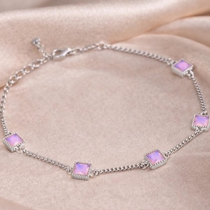 May include: A delicate silver bracelet featuring five square, light purple gemstones. The bracelet is displayed on a soft, light-colored fabric, highlighting the jewelry's elegant design and color.