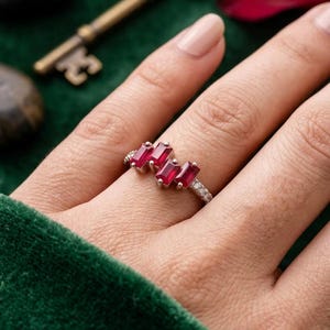 Könnte beinhalten: Ein silberner Ring mit vier im Baguetteschliff geschliffenen roten Rubinen, die in einer versetzten Gruppe angeordnet sind, akzentuiert durch kleine runde, klare Steine entlang der Ringschiene. Der Ring wird am Finger getragen und zeigt die roten Edelsteine.