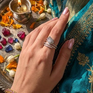 Könnte beinhalten: Ein silberner Ring mit einem blattförmigen Design, besetzt mit kleinen, funkelnden Steinen, wird an einem Finger getragen. Ein goldenes Armband mit roten Steinen ist ebenfalls zu sehen. Der Hintergrund umfasst einen türkisfarbenen Stoff mit Goldstickerei.