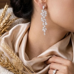 Könnte beinhalten: Elegante silberne Ohrringe mit einem kaskadierenden Blumendesign, das mit zahlreichen klaren, funkelnden Steinen besetzt ist. Ein passender Ring mit einem ähnlichen Blumencluster ist ebenfalls zu sehen. Der Schmuck ist vor einem Hintergrund aus Satin und weichem Strickstoff platziert.