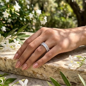 Könnte beinhalten: Nahaufnahme einer Hand, die einen silbernen Ring mit rechteckig geschliffenen Steinen trägt. Der Ring ist in einem Band aus kleinen, funkelnden Steinen gefasst. Im Hintergrund weiße Blüten und grünes Laub.