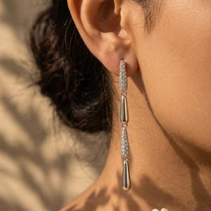 May include: Elegant silver drop earrings with sparkling accents. The earrings feature a linear design with three sections: two sections with small, glittering stones and a smooth, elongated bottom. A sophisticated jewelry piece.