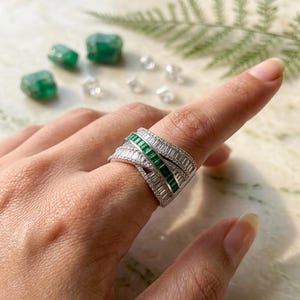 Könnte beinhalten: Ein silberner Ring mit einem geschichteten Design, der Reihen aus kleinen, klaren Steinen und ein zentrales Band aus rechteckigen smaragdgrünen Steinen aufweist. Der Ring befindet sich an einem Finger, mit anderen Smaragden und klaren Steinen im Hintergrund.
